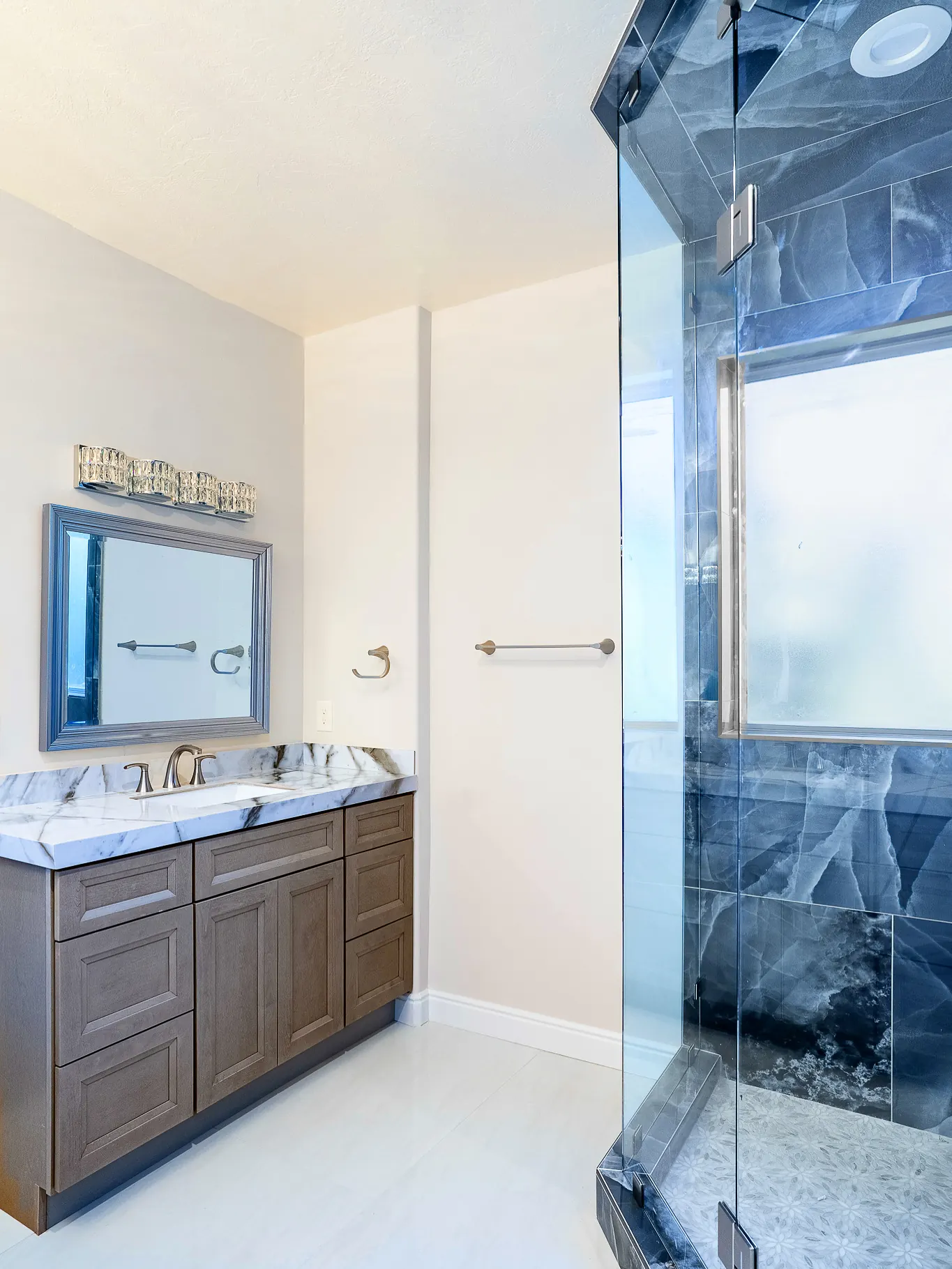Bathroom with a gray-brown vanity, white marble counter, and a glass shower lined in blue stone tile
