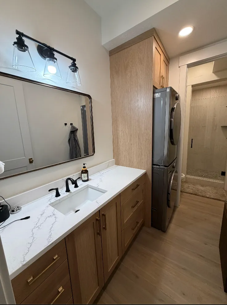 White-oak vanity with marble-look counter, matte-black faucets and mirror, stacked appliances beyond