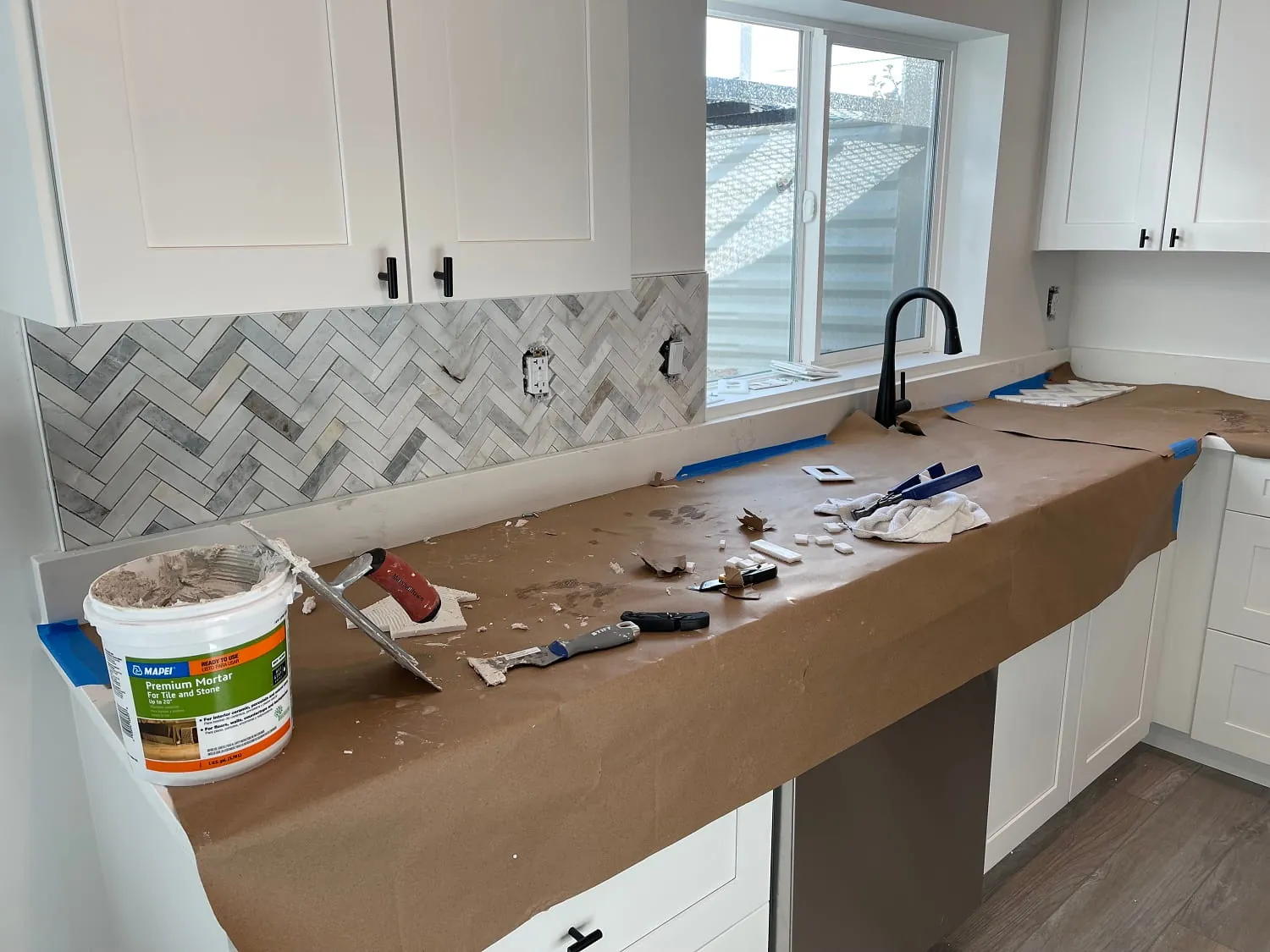 Herringbone backsplash install in progress at window/peninsula, thinset bucket on counter