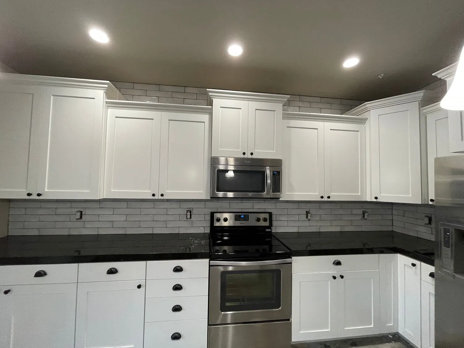 White shaker cabinets with black countertops, white subway backsplash, stainless range and microwave, recessed lights
