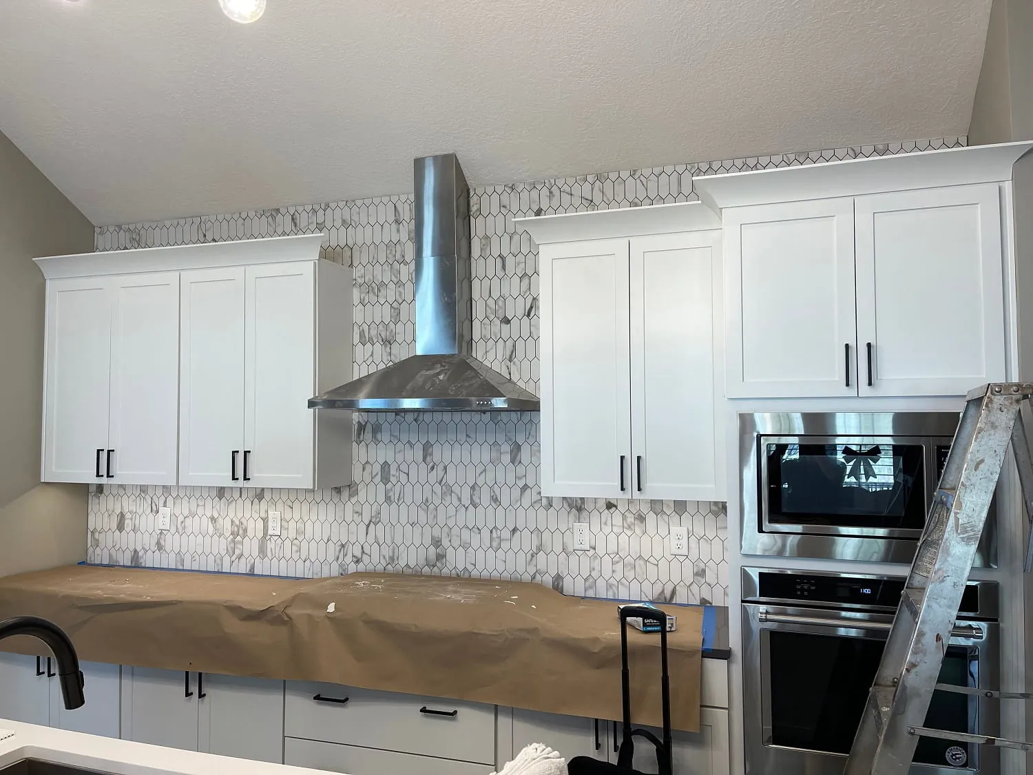 White shaker kitchen with stainless chimney hood, marble mosaic backsplash, double wall oven; counters still papered