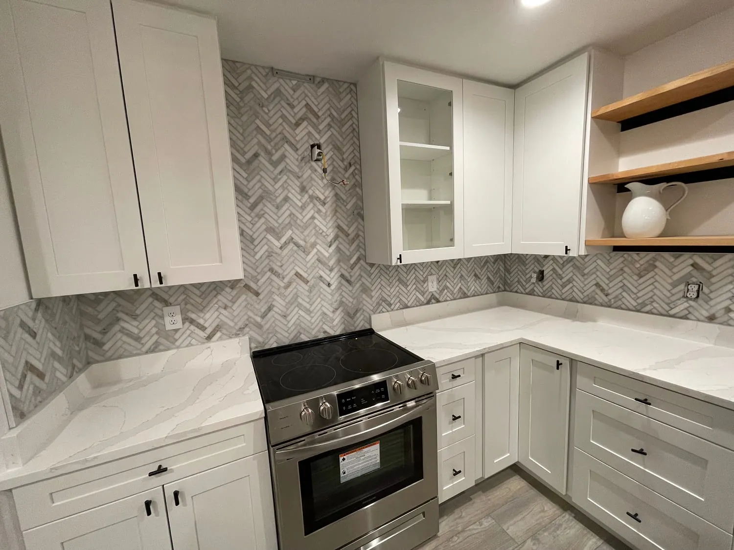 White shaker kitchen: marble herringbone backsplash, white quartz counters, floating wood shelves, stainless range