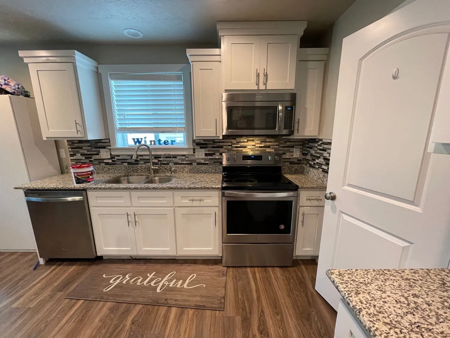 White shaker kitchen with window over sink, stainless appliances, granite/mosaic backsplash, wood-look floor